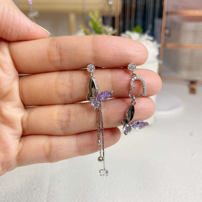 Pair of mismatched earring dangles in dream butterfly design crafted with semi-precious purple and colorless swarovski crystals, as well as a silver heart shape drops and two tassels drops, holding by a hand - Sisilia Jewels