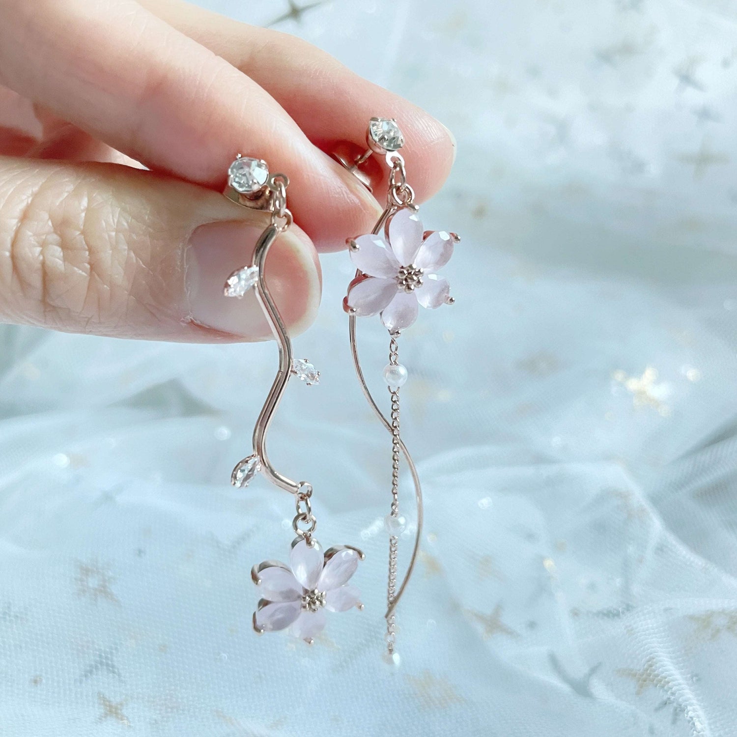 Mismatched earrings in six petals pink flower hanging on rose gold flower stems hold by three fingers - Sisilia Jewels